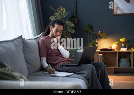 Young adult Asian man sitting on sofa working on laptop and writing notes in notebook, resting head on hand, appearing focused and thoughtful in modern living room setting Stock Photo