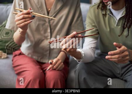 Asian woman and Black man sitting together holding chopsticks, demonstrating traditional eating technique, focusing on hands and upper bodies, sharing cultural experience indoors Stock Photo
