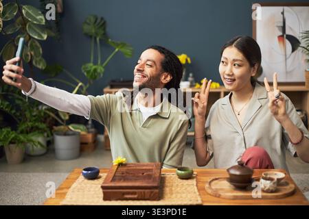 Young adult Black man taking selfie with smartphone while smiling next to young adult Asian woman making peace sign gesture, both sitting at table with traditional tea set Stock Photo