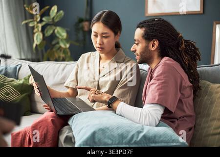 Asian young adult woman and Black young adult man sitting on sofa discussing information on laptop screen, both focused on device, collaborating in casual home setting Stock Photo