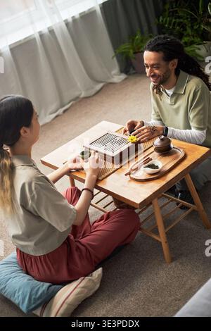 Asian woman and Black man sitting on floor playing traditional board game at low wooden table, both appearing as young adults, engaging in cultural activity with tea set visible Stock Photo