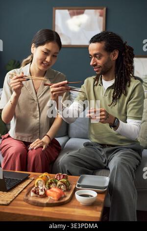 Asian young adult woman and Black young adult man sitting together holding chopsticks and sharing sushi, smiling and engaging in cultural dining experience in modern living room Stock Photo
