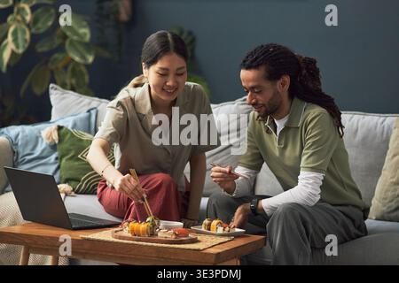 Asian young adult woman and Black young adult man sitting together eating sushi with chopsticks in living room, using laptop sharing meal and engaging in conversation Stock Photo