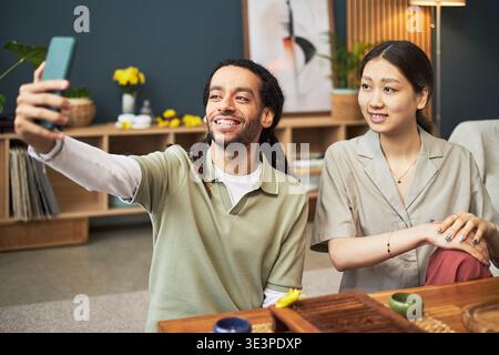 Young adult Black man smiling and holding smartphone taking selfie with young adult Asian woman sitting beside him, both engaging in casual conversation in modern indoor setting Stock Photo