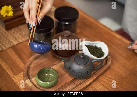Asian young adult woman preparing traditional tea ceremony using wooden chopsticks to handle ceramic teacup, tea leaves and teapot arranged on wooden tray reflecting cultural heritage Stock Photo