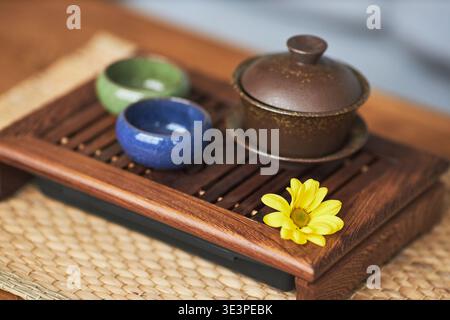 Traditional Asian tea set featuring ceramic teapot and two cups arranged on wooden tray with yellow flower accentuating cultural significance and ceremonial presentation Stock Photo