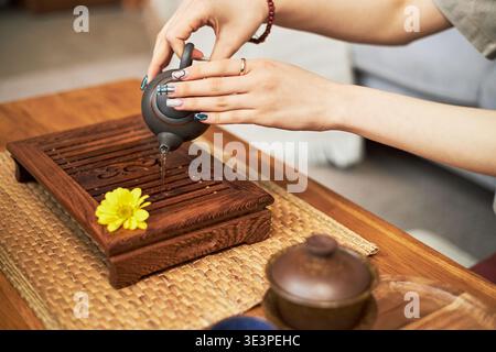 Asian young woman pouring tea from traditional teapot onto wooden tea tray during cultural tea ceremony, hands with manicured nails visible, yellow flower placed on tray Stock Photo
