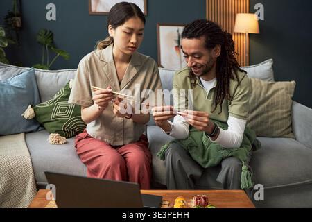 Asian young adult woman teaching Black young adult man using chopsticks while sitting together on sofa, both focusing on learning traditional eating technique with sushi on table Stock Photo