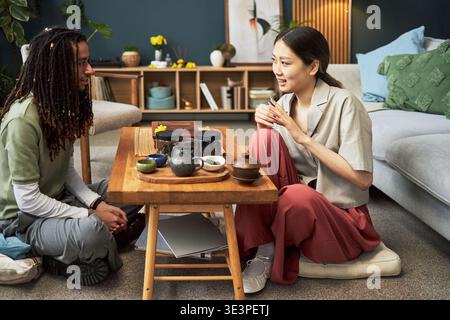 Young Asian woman and young Black man sitting on floor around low wooden table engaging in traditional tea ceremony, both smiling and interacting in modern living room setting Stock Photo