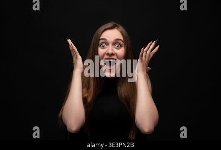 Young woman feeling surprised and excited, expressing shock and joy with wide eyes and open mouth on dark background Stock Photo