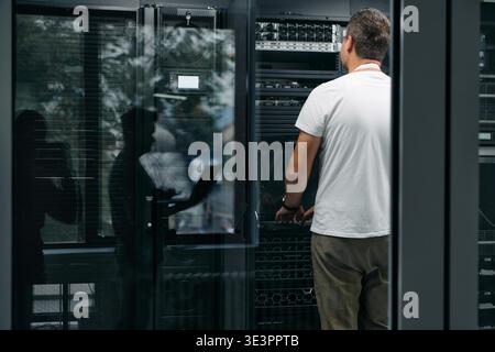 Data center technician inspects and maintains server racks and network hardware through a glass door, conveying professionalism, technical work and IT infrastructure maintenance in a controlled environment. || Model released Stock Photo