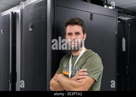 A confident data center technician stands with arms crossed beside black server racks, representing IT operations, network engineering, maintenance and secure enterprise technology in a modern server room. || Model released Stock Photo
