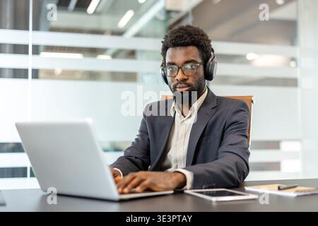 African businessman concentrating on his work, typing on a laptop while wearing headphones at a modern office desk, embodying professionalism, focus, Stock Photo