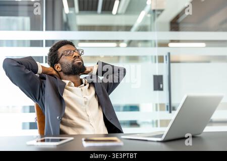 Confident young businessman leaning back in his office chair with hands behind his head and eyes closed, enjoying a calm, satisfied moment of rest aft Stock Photo