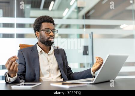 Thoughtful professional man with eyeglasses meditating at an office desk, finding focus and managing stress while sitting in front of a laptop and wor Stock Photo