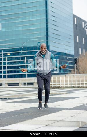 African american man jumping rope on plaza by glass office in sportswear with puddles Stock Photo