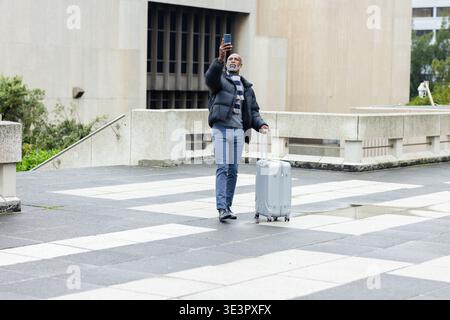 African american man standing in city square holding smartphone aloft and pulling rolling suitcase Stock Photo
