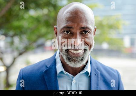 Senior african american man smiling outdoors beside tree and glass building in navy blazer Stock Photo
