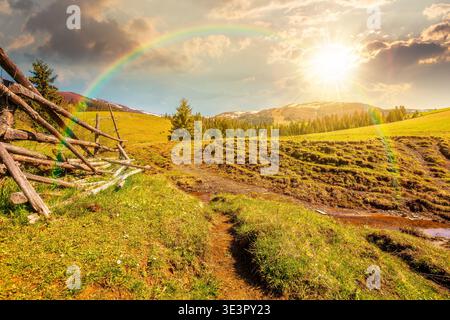 sunset over mountainous countryside landscape in spring. wooden fence and grassy meadow on a green hill. distant mountain ridge with snow capped tops. Stock Photo