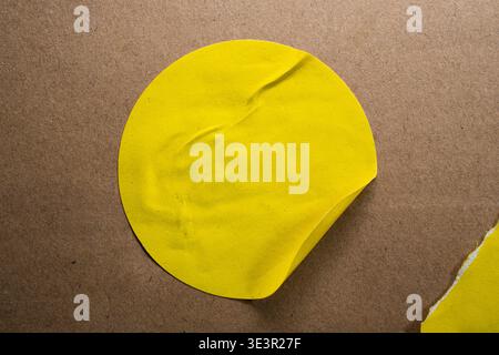 A close-up of a circular yellow paper sticker partially peeling from a smooth brown MDF wood board. The shot highlights the contrast between the vibra Stock Photo