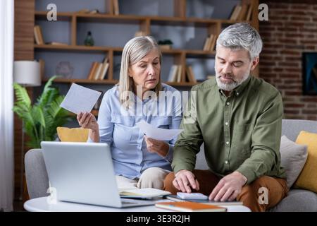 Senior couple sitting on a sofa at home, managing household budget, reading documents, using a laptop and calculator, planning their retirement financ Stock Photo