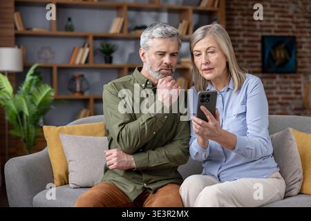 Senior couple on a living room sofa sharing a smartphone, looking thoughtfully at the screen as they discuss and navigate online information, technolo Stock Photo