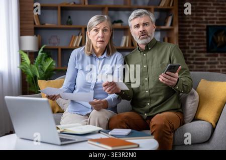 Senior couple sitting on a sofa in their living room, looking shocked and worried while reviewing financial documents, bills, and using a laptop for h Stock Photo