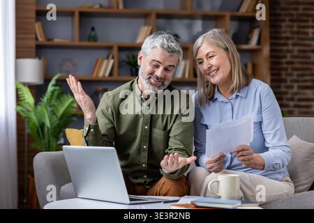 Mature husband and wife discussing financial planning and managing household budget, using a laptop and holding documents while relaxing on the couch Stock Photo