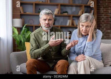 Mature couple having a serious disagreement on the couch, with the man expressing frustration while the woman appears upset and distressed, reflecting Stock Photo