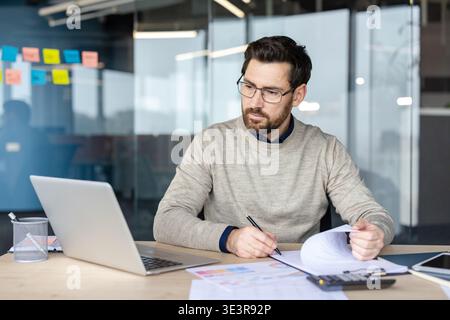 Professional man wearing glasses working on a laptop at his desk in a modern office, concentrating while reviewing financial documents, project schedules, and data analytics paperwork Stock Photo