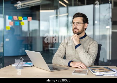 Man sitting at a modern office desk, resting his chin on his hand, engrossed in deep thought while planning future business strategies and solving problems Stock Photo