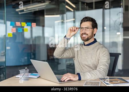 Happy business man sitting at modern workplace, adjusting his eyeglasses, looking away and smiling while typing on laptop during a successful day at work Stock Photo