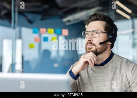 Bearded call center agent with glasses and headset in a modern office, looking away thoughtfully while providing customer support and technical assistance with concentration and professionalism Stock Photo