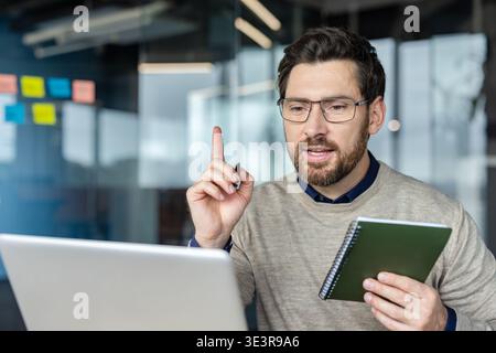 Man wearing glasses actively participating in online video conference, gesturing with pen while holding a notepad and explaining a new concept from his office desk Stock Photo