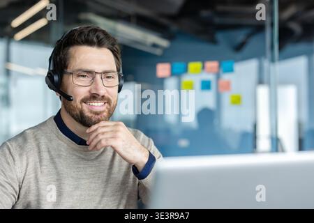 Bearded man with glasses wearing a professional headset and smiling at a laptop while providing online customer support or joining a virtual meeting, in a modern office Stock Photo
