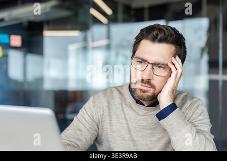 Focused businessman pondering a problem, working on his laptop in a modern office, embodying concentration, stress, and careful consideration for work and tasks Stock Photo
