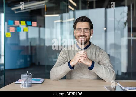 Confident male professional smiling while sitting at his modern office desk, representing business success, leadership, and a positive corporate environment Stock Photo
