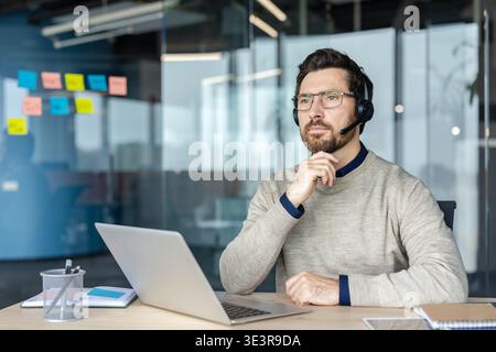Man wearing an online support headset, concentrating and thinking while working on his laptop at a modern office desk, providing communication and professional consultation Stock Photo