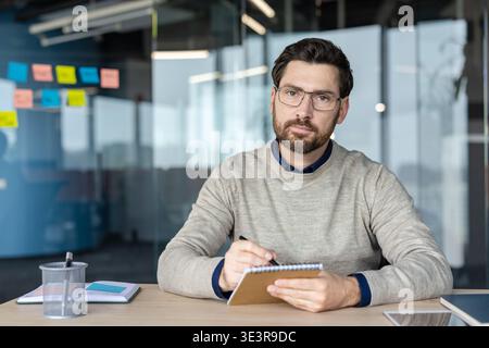 Businessman wearing glasses and a sweater sitting at a desk in a modern office, taking notes in a notebook, looking at the camera during a remote online video call Stock Photo