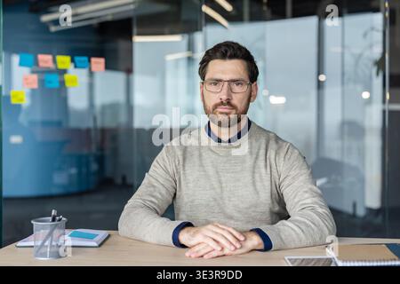 Professional man wearing glasses sitting attentively at a modern office desk against a blurred background of glass walls with sticky notes, representing a focused work environment Stock Photo