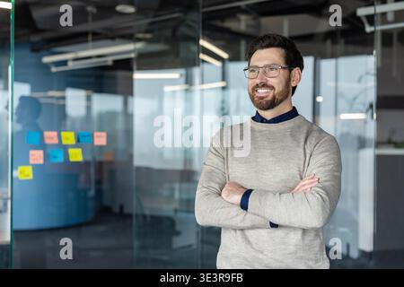 Smiling businessman standing with arms crossed in a contemporary glass office, looking away and projecting confidence, leadership, and professional success while planning new business strategies Stock Photo