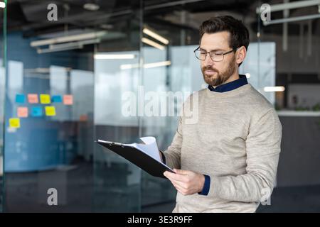 Professional man wearing glasses and a beard, focusing intently while reviewing documents on a clipboard, standing in a modern office with glass walls and colorful sticky notes Stock Photo