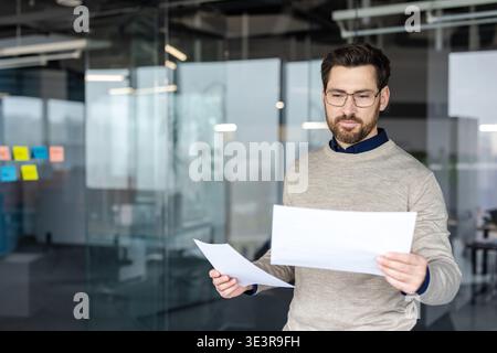 Young businessman standing in a contemporary glass office, reading and analyzing financial documents with a focused expression, representing data review and corporate management Stock Photo