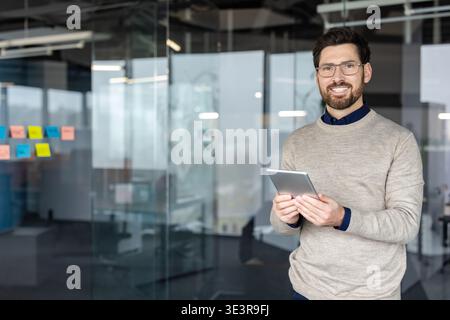 Young man smiling at camera, holding digital tablet in hands, standing in a contemporary workplace with glass walls and colorful sticky notes, representing modern business communication and success Stock Photo