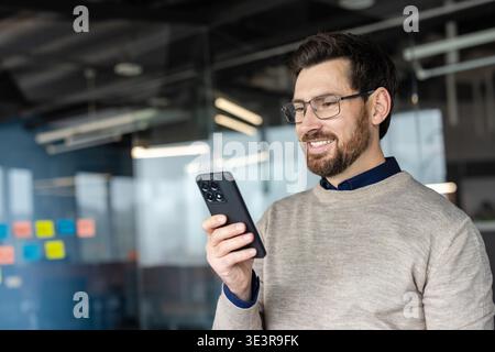 Bearded man in glasses and a sweater smiling while using a smartphone, standing in a modern corporate office and scrolling through messages and apps while engaging with coworkers Stock Photo