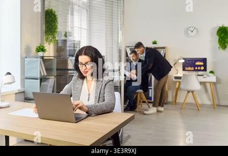 Businesswoman working on laptop in office while colleagues discuss financial report Stock Photo