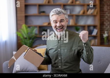 Excited senior man is holding an open cardboard box, showing joy and satisfaction after receiving a parcel from an online order while sitting on a couch at home Stock Photo