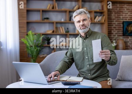 Mature man at home office reviewing receipts and documents with laptop, calculator and notebook, focused on budgeting, expenses and personal or small business finances Stock Photo