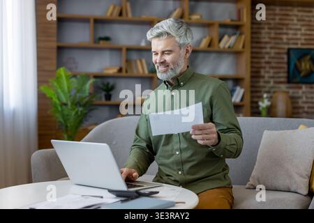 Mature man with gray hair and beard using laptop on sofa, reviewing documents and managing online banking, bills, retirement savings and household finances at home Stock Photo