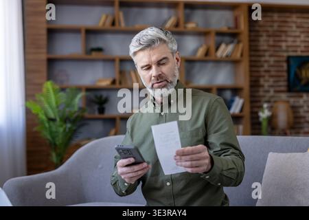 Mature man on sofa at home comparing paper receipt with smartphone, managing household finances and paying bills online with banking app, focused and responsible Stock Photo